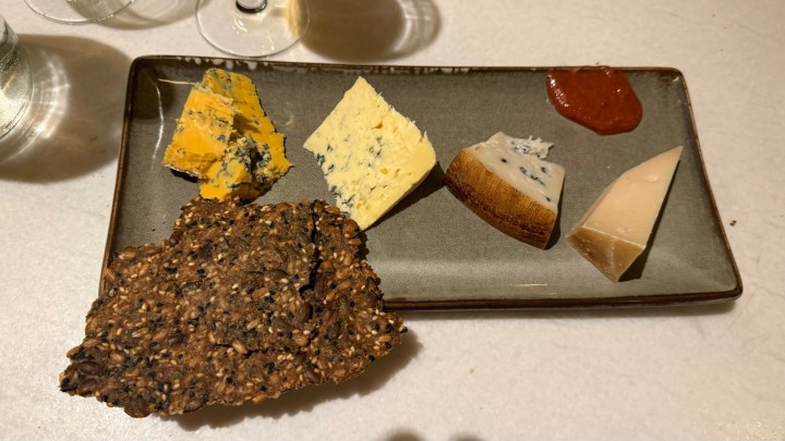 A plate featuring an assortment of five cheeses, including yellow and blue varieties, accompanied by a piece of dark grain cracker and a small dollop of red sauce.