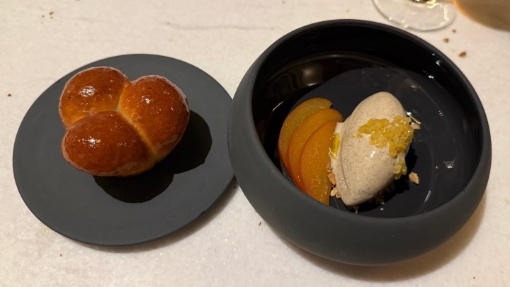 A glazed brioche bun shaped like a heart on a black plate next to a round black dish containing a scoop of ice cream topped with crushed nuts and sliced fruit.