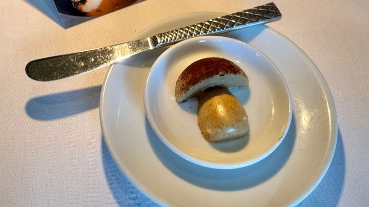 A creatively designed dessert resembling a mushroom, placed on a small white plate with a stainless steel knife beside it.