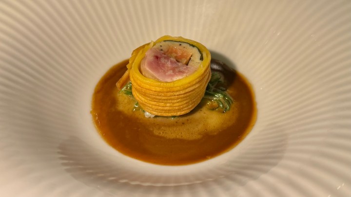 A gourmet dish featuring a layered cylindrical presentation of yellow pasta or crepe, filled with seafood and greens, served in a rich brown sauce.