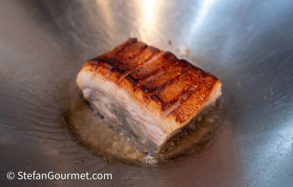 A piece of crispy-skinned pork belly cooking in a metal pan with a sheen of rendered fat.