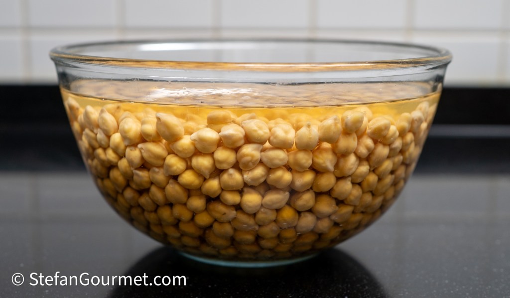 A clear glass bowl filled with soaked chickpeas in water.