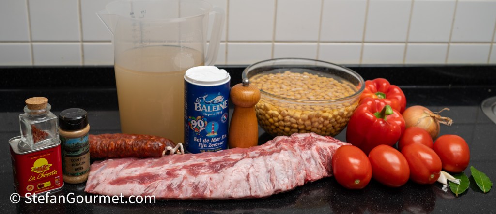A spread of ingredients for a recipe, including a slab of meat, spices, a jar of chickpeas in water, red bell peppers, fresh tomatoes, garlic, onions, bay leaves, and broth in a measuring cup.