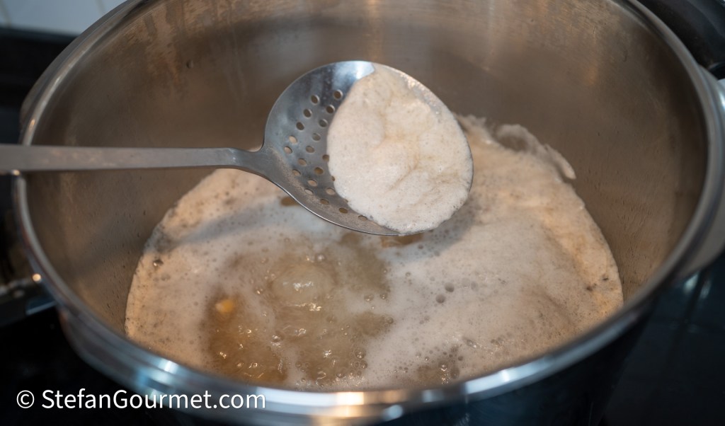 A metal ladle scooping frothy liquid from a pot on a stovetop, with bubbles and steam visible in the pot.