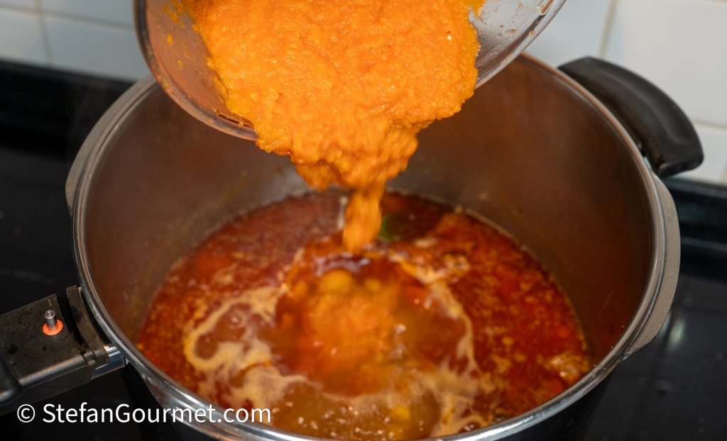 A pot on a stove with a thick orange sauce being poured into it from a ladle.