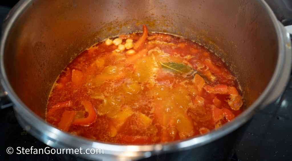 A pot of simmering soup with colorful vegetables including red bell peppers, chunks of carrots, and spices, on a stovetop.