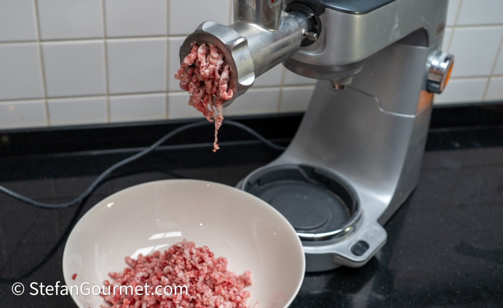 Freshly ground meat being extruded from a meat grinder into a white bowl, with a kitchen aid mixer in the background.