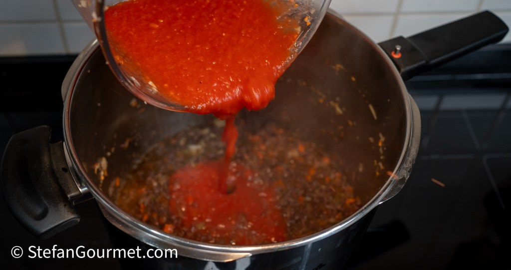 A person pouring red sauce from a measuring cup into a pot containing cooked ingredients, with steam rising from the pot.