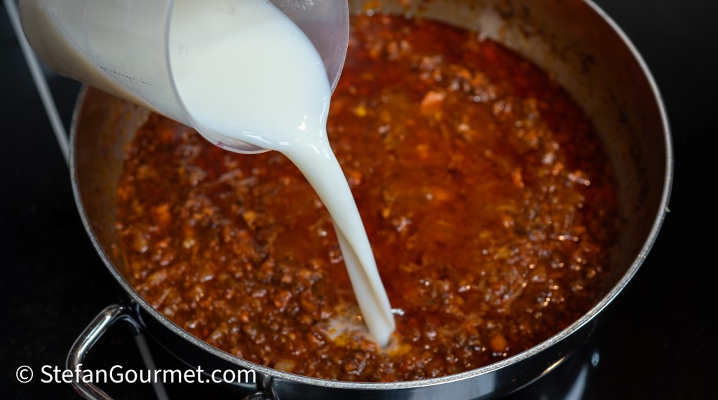 A person pouring milk from a jug into a pot of simmering sauce.