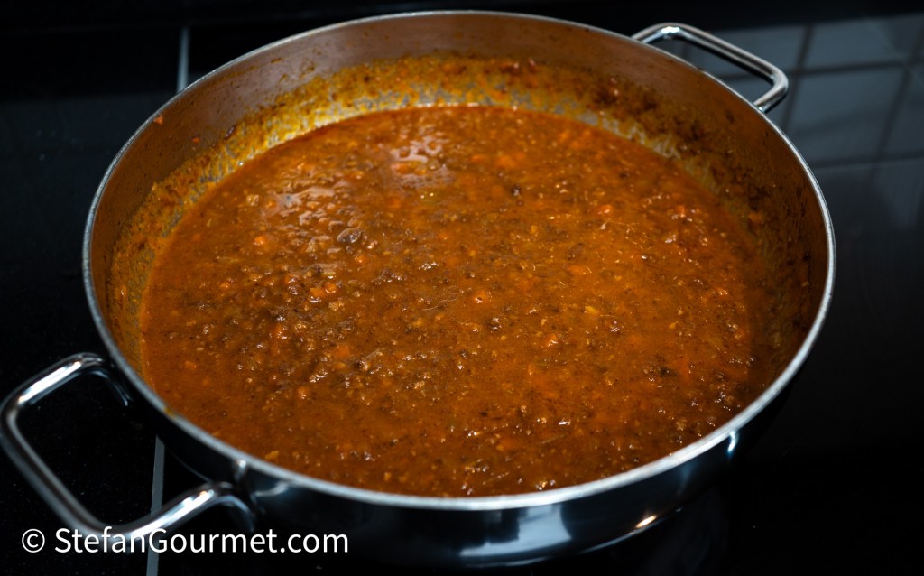 A large pot of simmering brown meat sauce, with visible herbs and spices, on a stovetop.