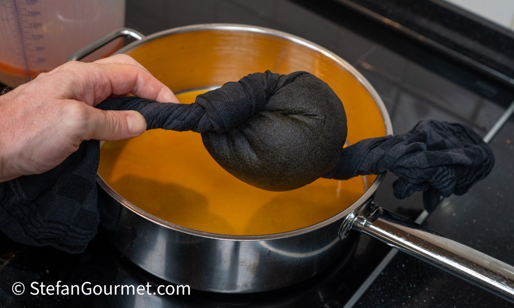 A hand holding a tied black cloth pouch, partially submerged in a pot of orange liquid on a stovetop.