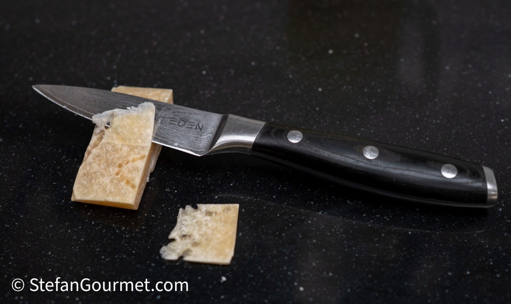 A close-up of a sharp kitchen knife resting on a black countertop, with two small pieces of cheese beside it.