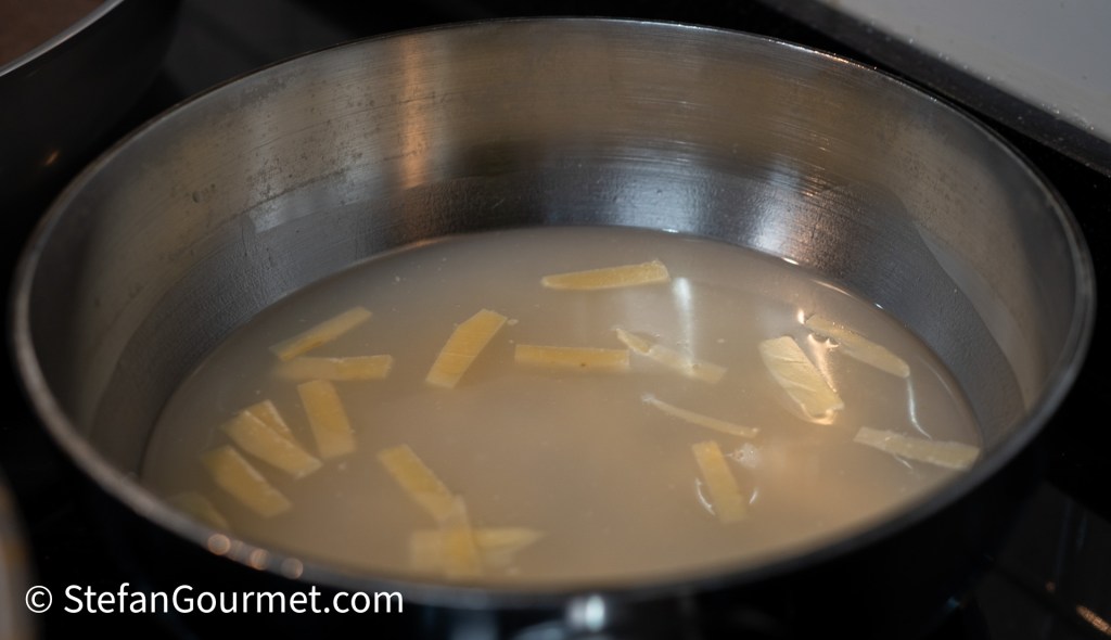 A metal saucepan filled with clear liquid and strips of pasta, simmering on a stove.