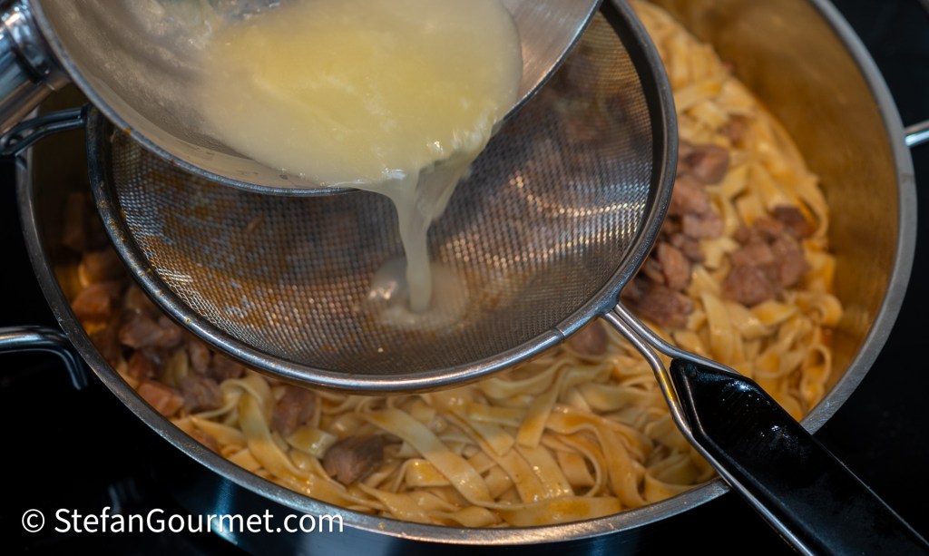 A close-up of a colander pouring liquid over cooked fettuccine pasta and meat in a pot.