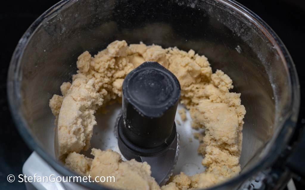 Close-up of a food processor containing a mixture resembling dough or cookie batter, with a central blade and partially formed consistency.
