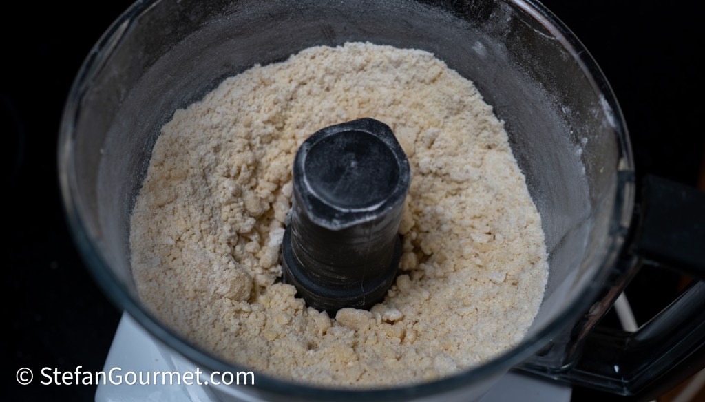 Close-up of ground flour or meal inside a food processor bowl, featuring a black processing blade in the center.