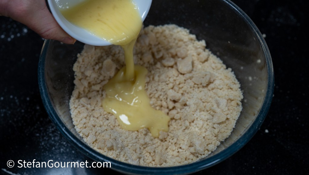 A hand pouring liquid mixture into a bowl of crumbly ingredients.