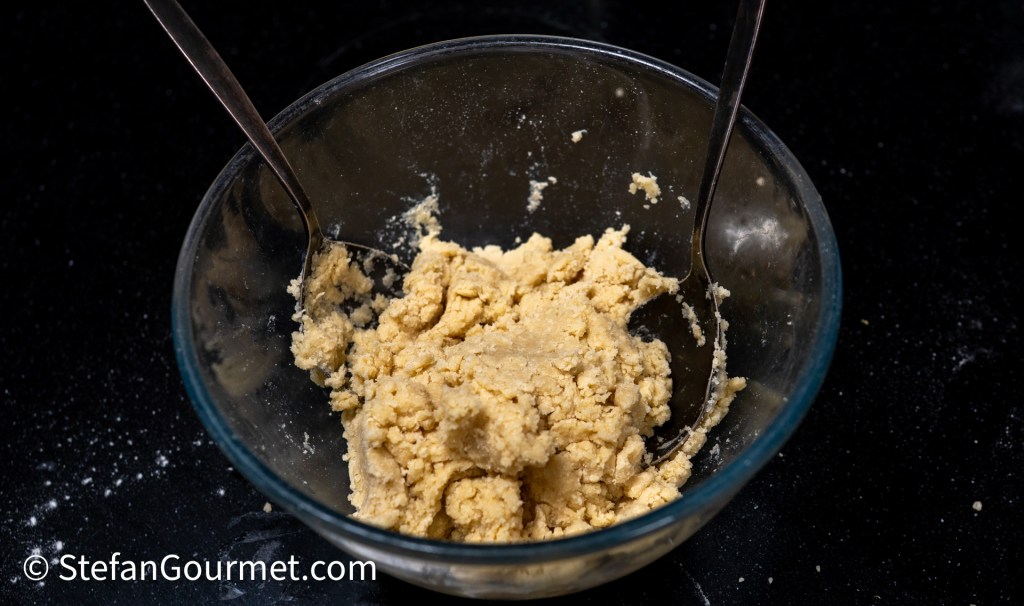 A glass bowl containing a thick, crumbly mixture, with two metal spoons resting inside. The bowl is placed on a dark surface, and some flour particles are scattered around.