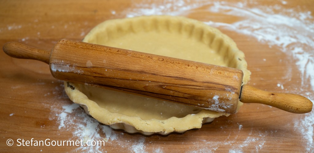 A wooden rolling pin rests on top of an unbaked pie crust in a pie dish, surrounded by flour on a wooden countertop.