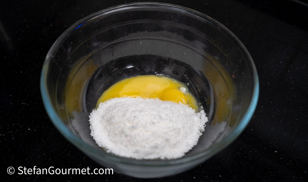 A glass bowl containing an egg yolk and granulated sugar on a dark countertop.