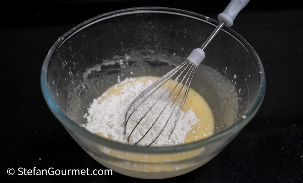 A glass bowl containing a mixture of batter and flour, with a whisk resting inside.
