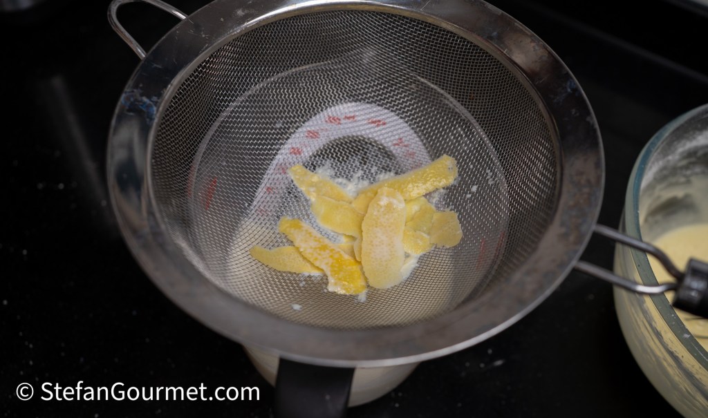 A fine mesh strainer containing strips of candied ginger and powdered sugar, positioned over a bowl with a creamy mixture.