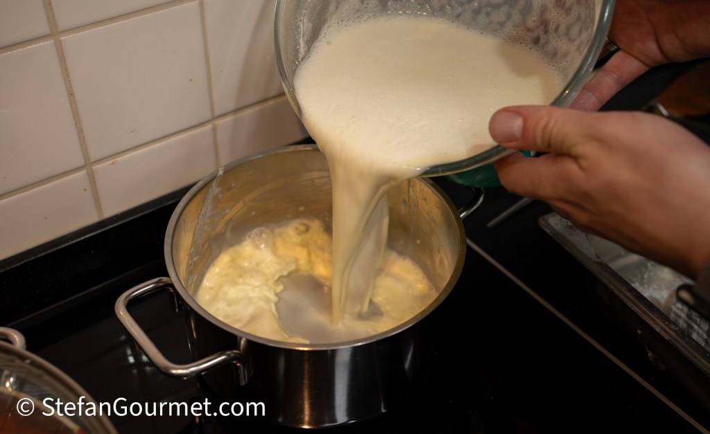 A person pouring a creamy mixture from a glass bowl into a stainless steel pot on a stove.