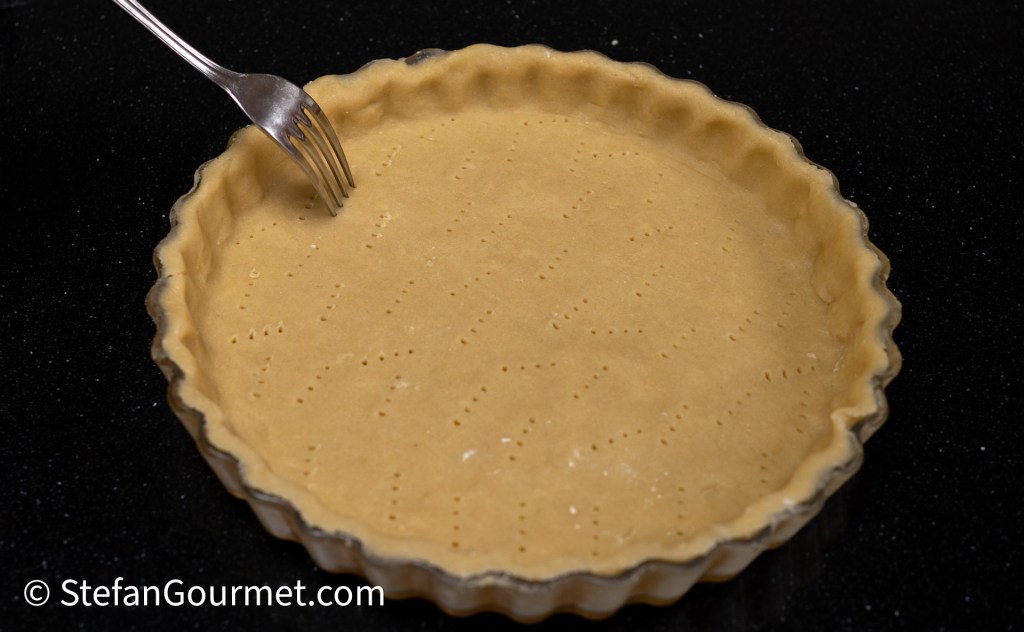 A partially baked tart crust with fork pricks to prevent bubbling, set on a black countertop.