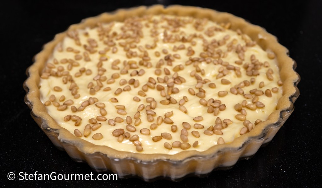 A freshly baked pie with a creamy filling topped with scattered pine nuts in a clear glass pie dish.