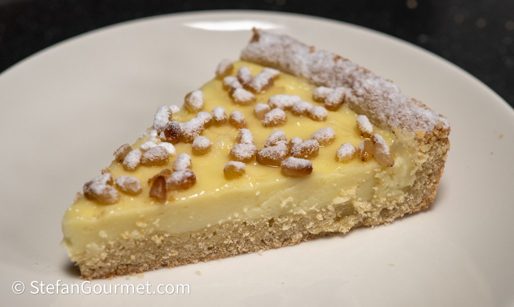 A slice of creamy lemon tart topped with toasted pine nuts and powdered sugar, served on a white plate.