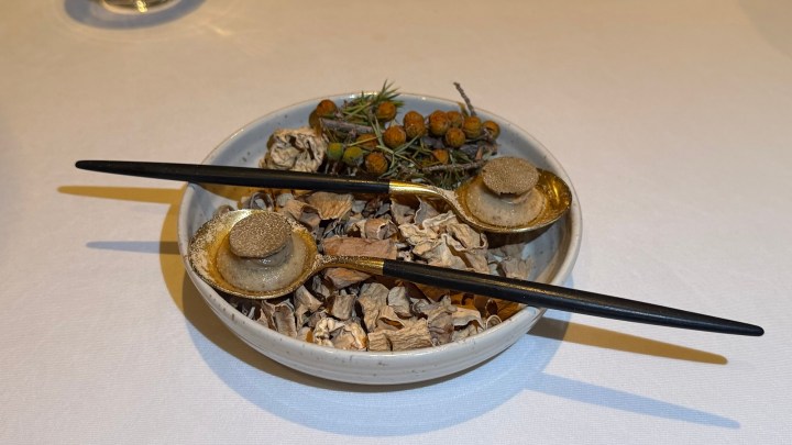 A ceramic bowl filled with dried leaves and herbs, featuring two gold spoons with round, textured items on top, and some green twigs and orange berries placed beside them.