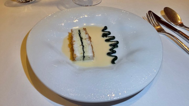 A elegantly plated dish featuring a rectangular slice of cake layered with cream and green garnish, accompanied by a drizzle of sauce on a white plate.