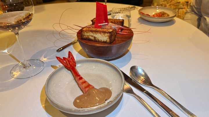 A gourmet dish featuring a large shrimp on a plate with a creamy sauce, alongside a bowl containing toasted pieces of bread. A glass of rosé wine and utensils are visible on the table.