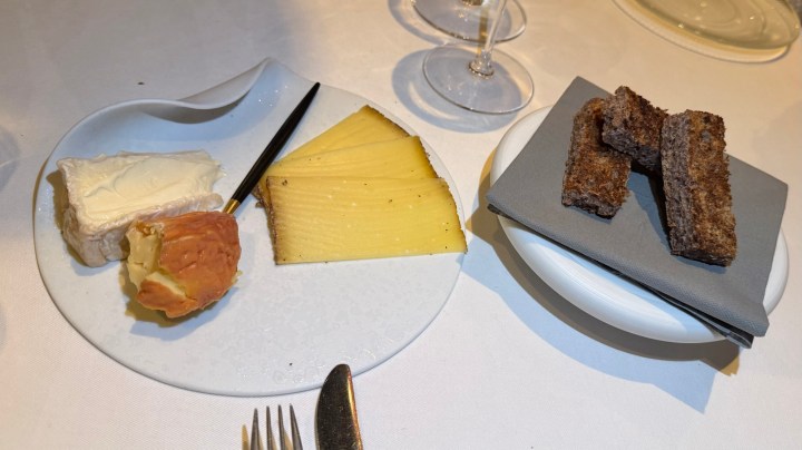 A plate with various cheeses, including a creamy cheese, a wedge of aged cheese, and small pieces of dark bread, accompanied by a napkin and wine glasses in the background.
