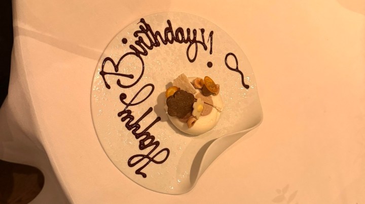 A dessert plate with the words 'Happy Birthday!' written in chocolate sauce, featuring a small cake topped with seasonal fruits and decorative elements.